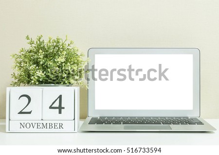 White wooden calendar with black 24 november word with white blank space at center of computer notebook on white wood desk and cream wallpaper textured background , selective focus at the calendar