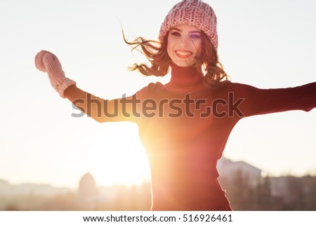 Winter portrait of a young smiling woman in a pink hat and mittens on a background sunset