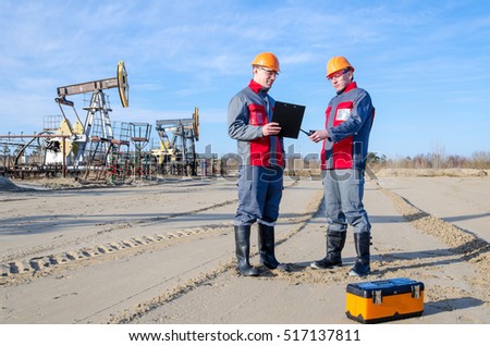 Two workers in the oilfield, one holding the radio second showing papers. Pump jack and wellhead background. Oil and gas concept. 