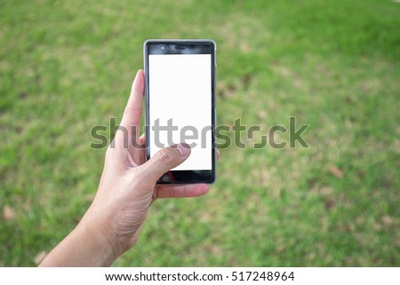 Man's left hand hold blank white screen mobile phone with blurry scene of green grass in park.