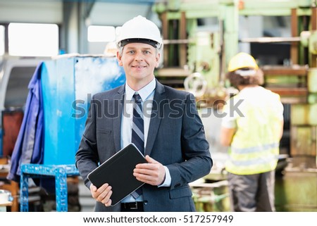 Portrait of confident mature businessman holding digital tablet with worker in background at factory