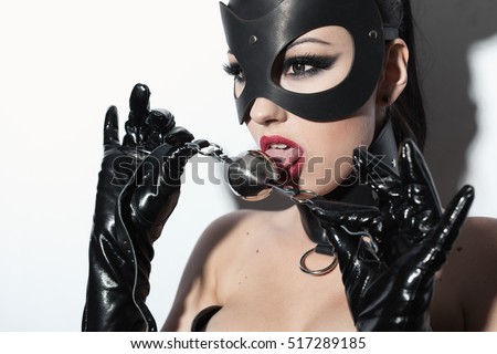 Beautiful dominant brunette vamp mistress girl in latex corset, long gloves, collar and bdsm black leather fetish mouse mask posing with metal ball gag on white backgroung Royalty-Free Stock Photo #517289185