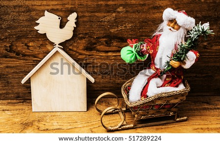 Merry Christmas! wooden rooster sit on roof of house. Card in cartoon style. Santa Claus with gift sack and green christmas tree sitting in sleigh on wooden wall background 