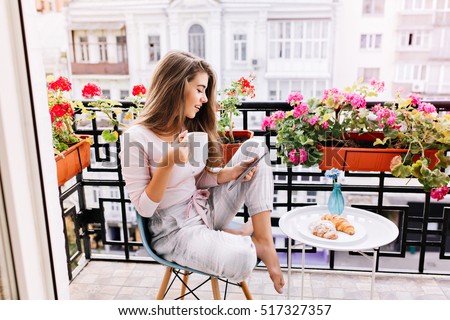 Attractive girl with long hair in pajama having breakfast on balcony in the morning in city. She holds a cup, reading on tablet Royalty-Free Stock Photo #517327357