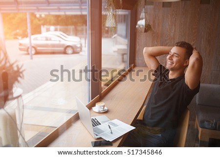 Happy day dreamer. Relaxed young man holding hands behind head and smiling while sitting at the table with laptop in modern cafe Royalty-Free Stock Photo #517345846