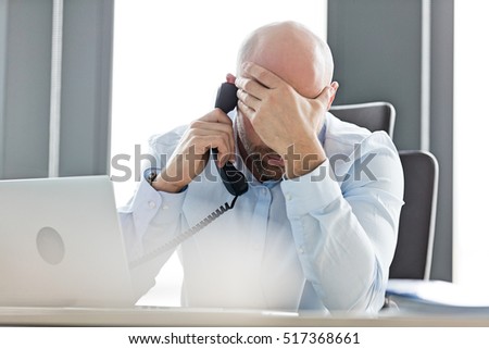 Tired mid adult businessman using landline phone at desk in office