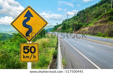 Warning traffic sign on metal pole winding road on background