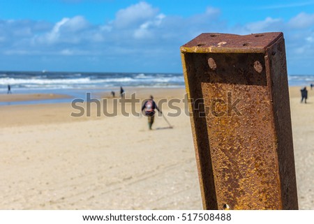 Rusty pole on the beach, guy with metal detector in the blurry background, walking over the north sea beach in northern holland. Travel photo.