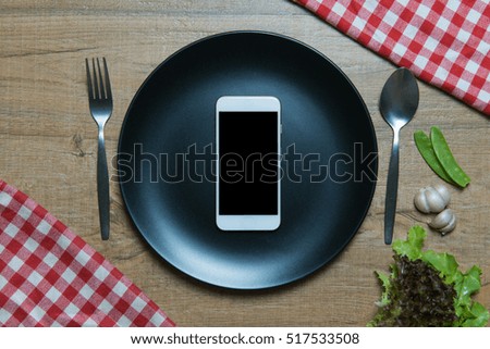 Top view of smartphone in empty black plate on wooden table and some green vegetable.