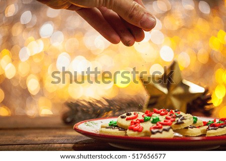 Organic Gluten Free Smiling Gingerbread Sugar Cookies On Holiday Plate And Wooden Background By Glowing Light Fireplace With Hand Dropping Sprinkles On Top.