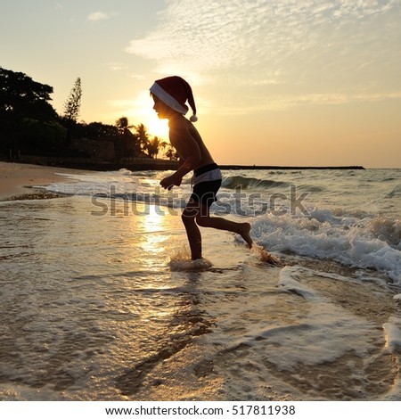 A square picture: Cute boy in a Santas cap standing at the sea playing and jumping. Against the background of the sun goes down, wonderful golden sheen on the sea surface