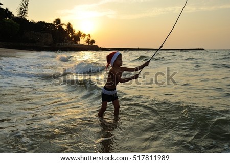 Cute boy in shorts and a Santas cap standing knee-deep in the sea and throws the bait, waiting for the catch. Against the background of the sun goes down, wonderful golden sheen on the sea surface