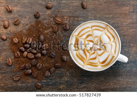 Hot Cappuccino coffee with zigzag caramel motif or spiderweb art floating on top and coffee beans with ground on wooden background. Coffee break at retro style coffee shop,t op view. Royalty-Free Stock Photo #517827439
