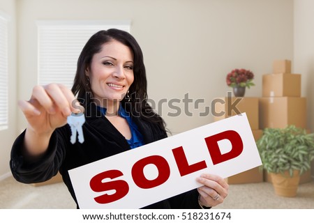 Happy Hispanic Female Real Estate Agent with Sold Sign and Keys in Room with Moving Boxes.