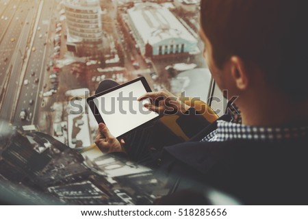 Mock up template: digital tablet in hands of businessman sitting near window of skyscraper and using modern tablet pc with empty screen for message or your logo, winter cityscape and highway outside