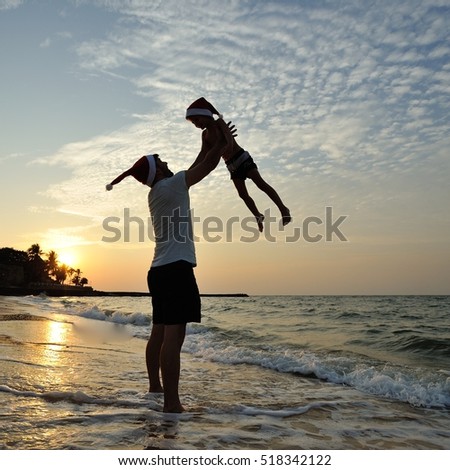 A square picture: silhouettes on the beach with a background of a golden sunset: dad throws up overhead his five year old son. On the heads of both Santaâ??s red cap, they are laughing