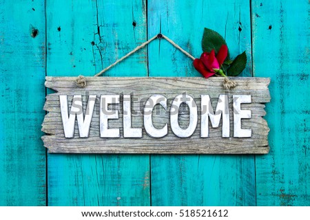 Weathered welcome sign with red rose bud hanging on antique rustic teal blue wood door; holiday background with home concept