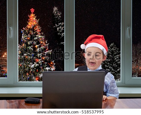 In cap of Santa Claus boy looking at a laptop on the background of the Christmas tree outside the window
