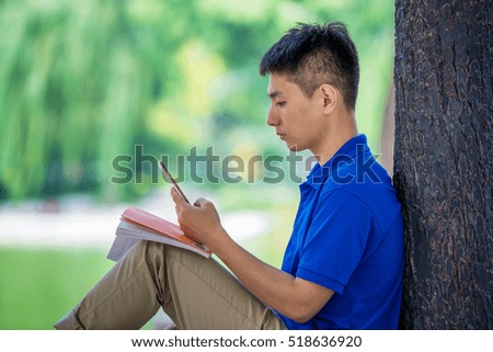 Young handsome Asian man enjoying his smart phone in public park sitting on bench and leaning trunk of a tree