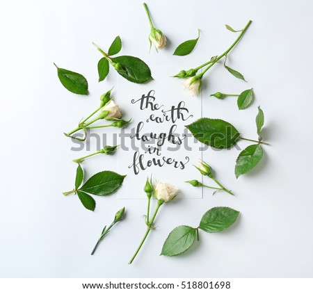 Quote "The earth laugh in flowers" written on paper with roses and leaves on white background. Top view