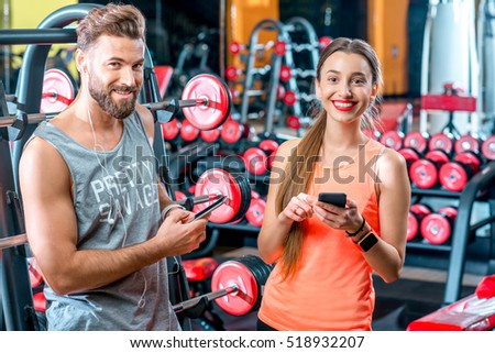 Sports couple standing with smart phones resting after the training in the gym with red dumbbells on the background