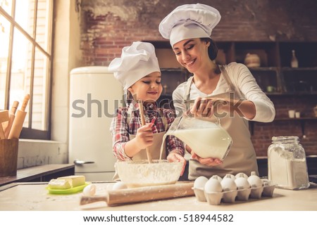 Cute little girl and her beautiful mother in chef hats are mixing muffin batter in the bowl and smiling while preparing it for baking. Mother pouring milk Royalty-Free Stock Photo #518944582
