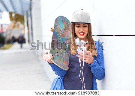 Young girl with skateboard leaning on modern gray wall. She is looking at mobile phone.