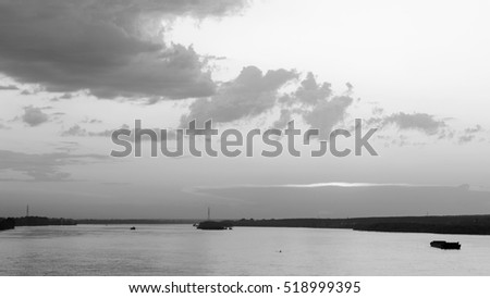 Black-and-white photograph of the Ob river with the sun setting through a gap in the clouds and the shadow of the floating barges.