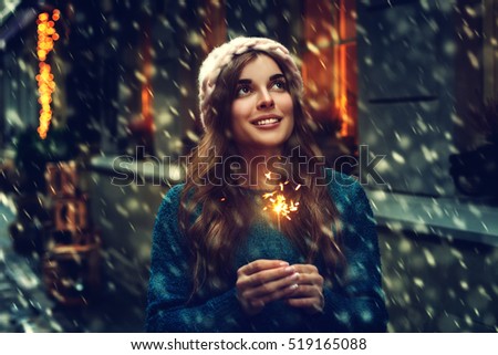 Outdoor photo of young beautiful happy smiling girl holding sparkler, walking on street. Model looking up, wearing stylish winter clothes. Waist up. Christmas, New Year, concept. Magic snowfall. Toned