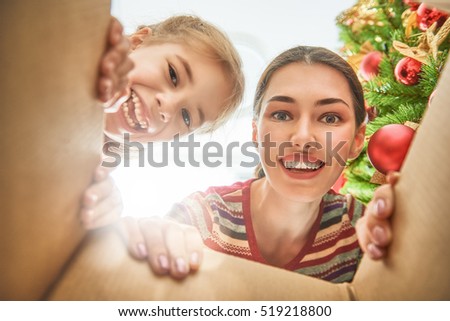 Merry Christmas and Happy Holidays! Cheerful mom and her cute daughter girl opening a Christmas present. Parent and little child having fun near Christmas tree indoors. View from inside of the box.