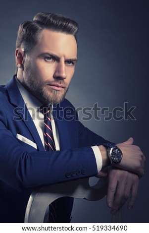 Young handsome bearded caucasian man with blue eyes sitting on chair. Perfect skin and hairstyle. Wearing blue suit and tie. Studio portrait on gradient black to grey background. Toned