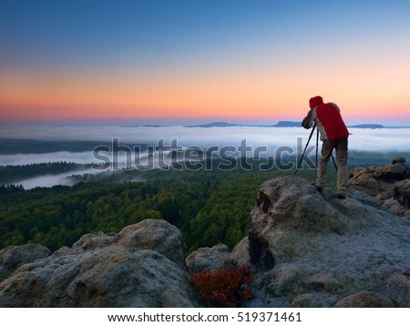 Photographer takes photos with mirror camera and tripod on peak of rock. Fall fogy landscape, spring orange pink misty sunrise in beautiful valley below.