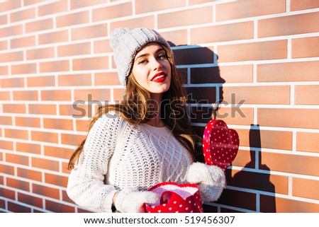 Portrait young girl with long hair in knitted hat, warm sweater and gloves on wall background outside. She holds open box heart in hands, smiling to camera