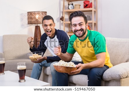 Picture of happy friends drinking beer and eating pop corn at home while watching football on TV. Happy handsome men posing for photographer.