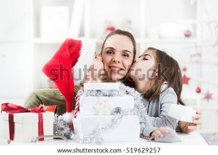 Two kids and their mother are opening Christmas presents in their living room. Kids are kissing their mother