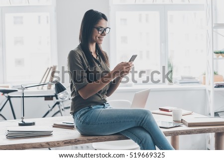 Typing business message. Confident young woman in smart casual wear holding smart phone and looking at it with smile while standing near her working place Royalty-Free Stock Photo #519669325