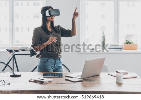 Innovation in business. Confident young woman in virtual reality headset pointing in the air while standing near her working place in office Royalty-Free Stock Photo #519669418
