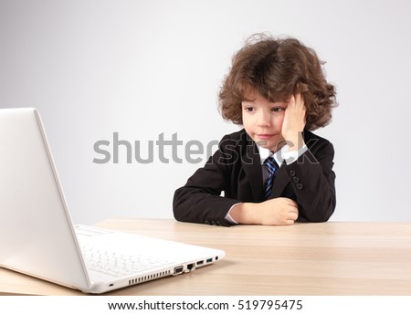 Little curly-haired businessman sitting at the table and looking at a laptop. Gray background.
