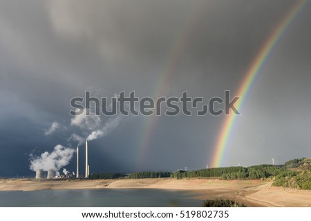 Double rainbow on stormy sky near coal power plant spewing smoke into the air.