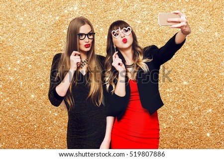 Two women having  great time together, making self portrait , use carnival funny paper glasses, wearing  elegant evening dress. Friends celebrating  new year or birthday party. 