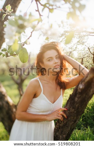 Young pretty serious caucasian woman in white dress standing near blooming spring tree. Youth, freshness, beauty, happiness concept. Tilt-shift image, intentionally blurred, soft selective focus.