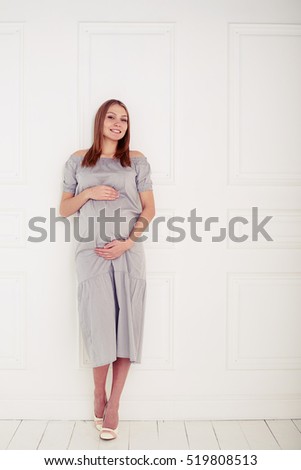 Mid shot of pregnant woman posing over white background. Holding her pregnant belly. Young smiling beautiful pregnant woman in grey loose dress