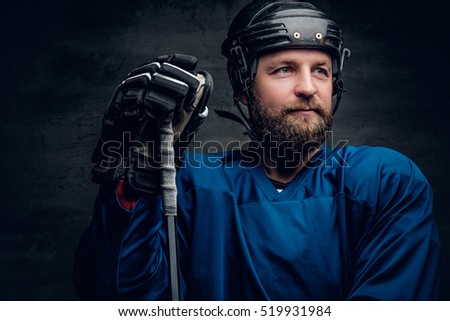 A bearded ice-hockey player in a blue sportswear holds a gaming stick in contrast illumination on grey vignette background.