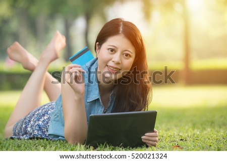 beautiful asian woman using digital tablet shopping online with credit card sitting on grass. outdoors background. technology and people concept