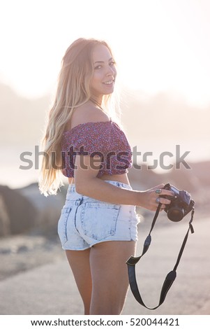 Pretty as a portrait. Young, attractive female looks over her shoulder and gives a cute smile. She is holding onto a camera and taking pictures. Focus is soft and dreamy.