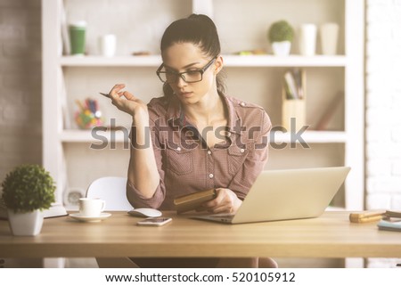 Cute caucasian female sitting at her office desktop with laptop, decorative plant and other items, looking away from the camera