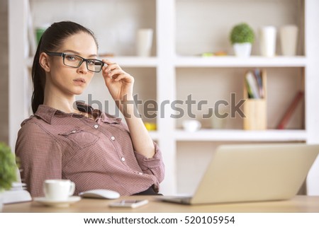 Cute caucasian woman sitting at her office desktop with laptop, decorative plant and other items, looking at the camera