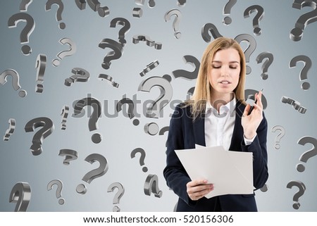 Portrait of a blond businesswoman with documents. She is standing near gray wall with question marks falling