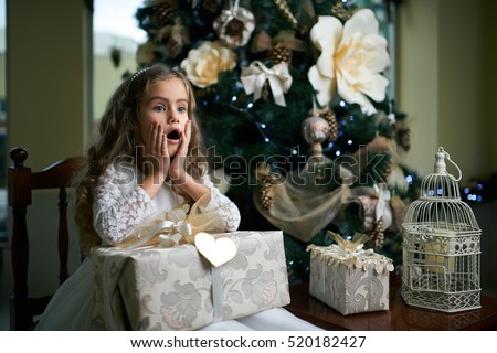 Little girl sits near Christmas tree with a gift and scared.