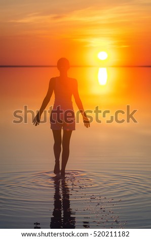 Beautiful female model open arms under the sunrise at seaside. Calm water of salt lake Elton reflects silhouette of woman. Sun goes under horizon. Girl stands alone in water. Sun reflects in water
